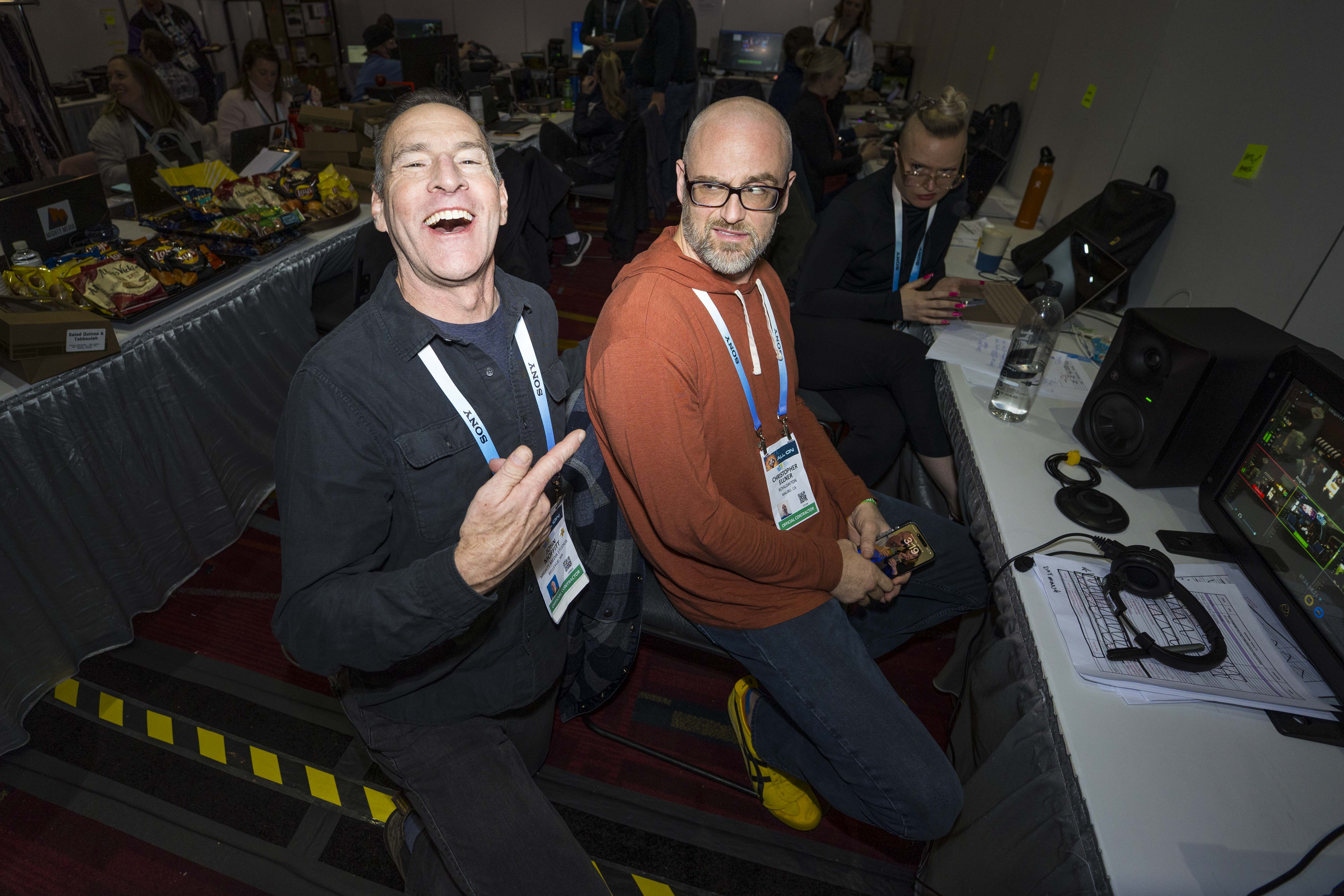 Three people at a control desk with monitors and headphones; one man laughs and points at another man.