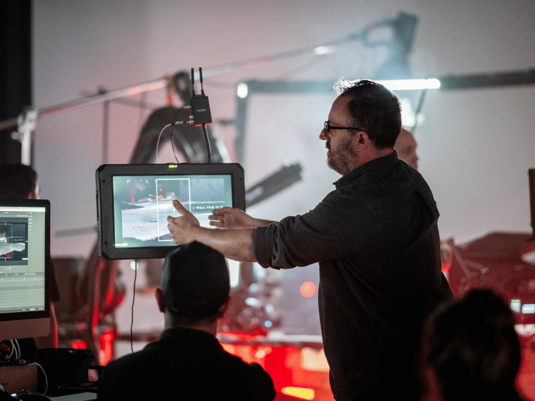 Man with glasses directing a video shoot, pointing at a screen on set with crew members around.