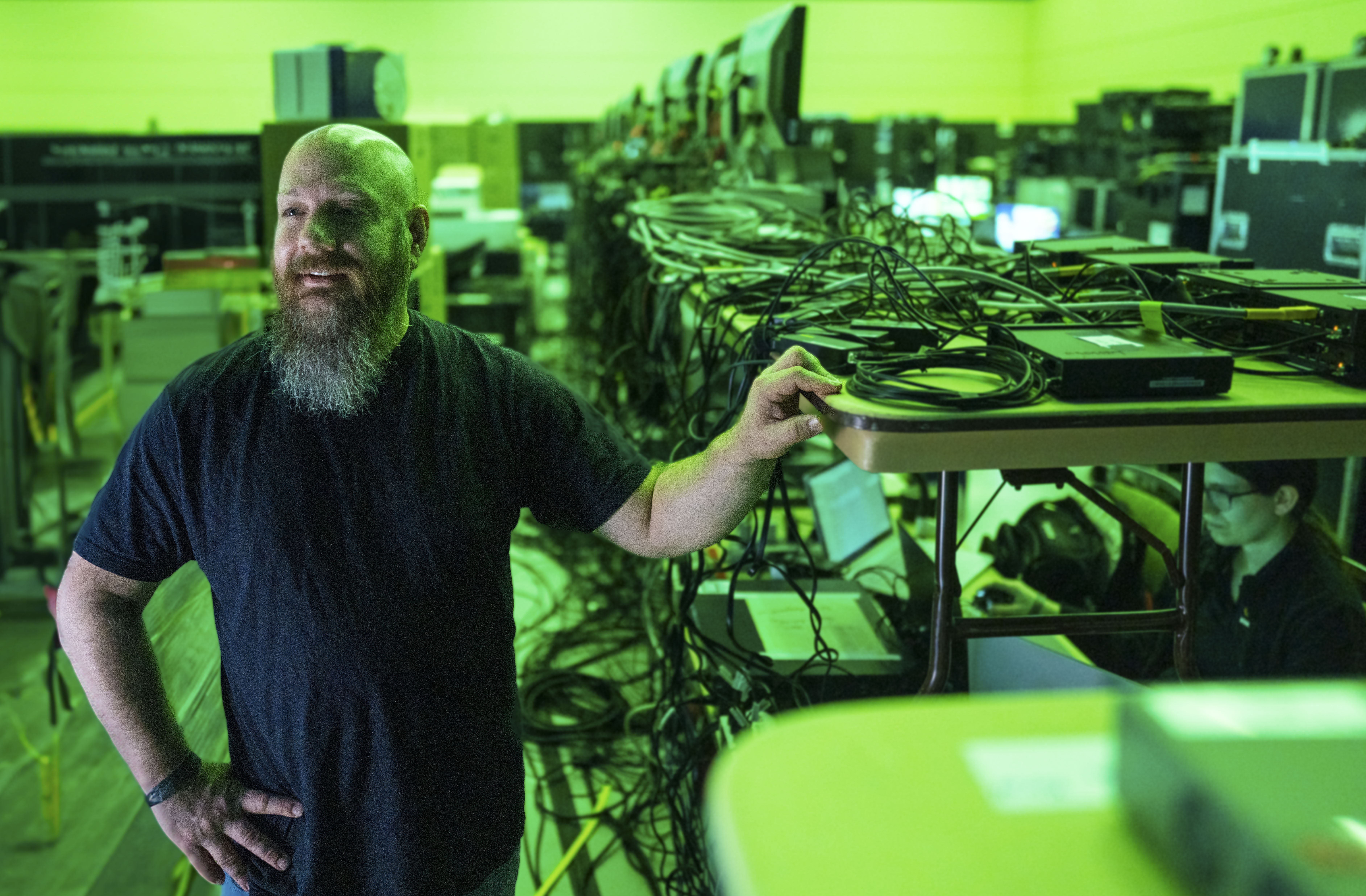 Bearded man stands by table with network devices and cables in a green-lit tech workspace.