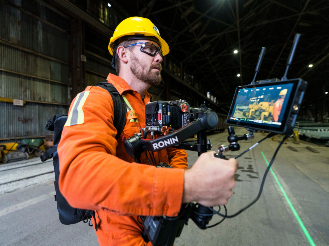 Man in orange workwear and yellow helmet filming inside industrial warehouse using camera stabilizer.