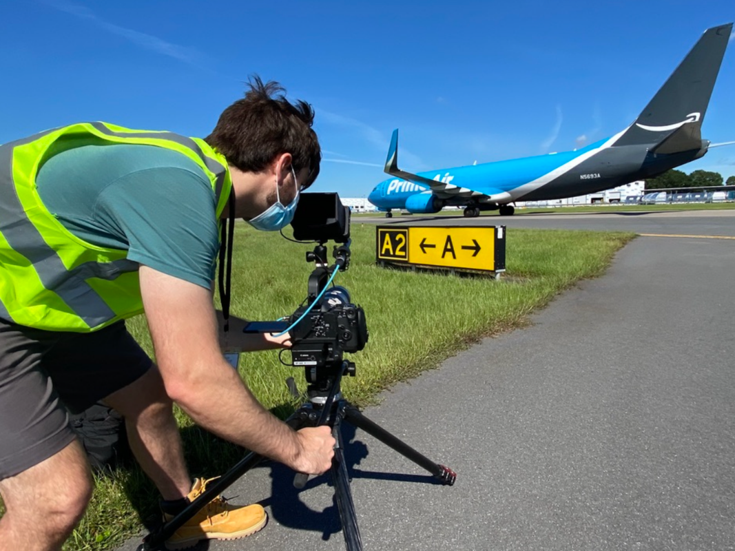 Person wearing mask and safety vest filming a blue Prime Air plane on a runway with a camera on a tripod.