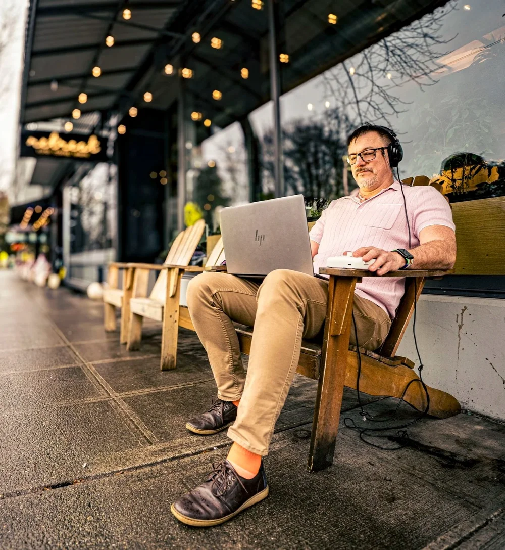 Man wearing headphones using a laptop while sitting on a wooden bench outside a cafe.