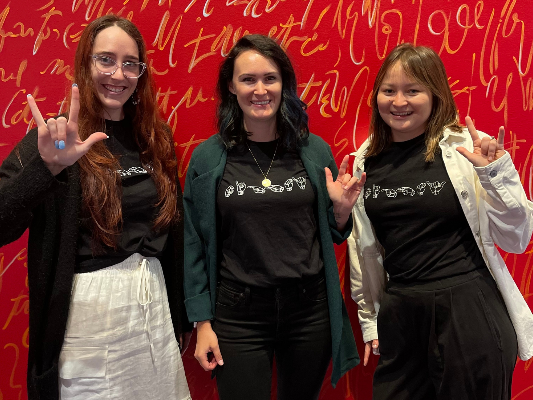 Three smiling women stand against a red wall, wearing black sign language t-shirts and showing the I love you ASL sign.