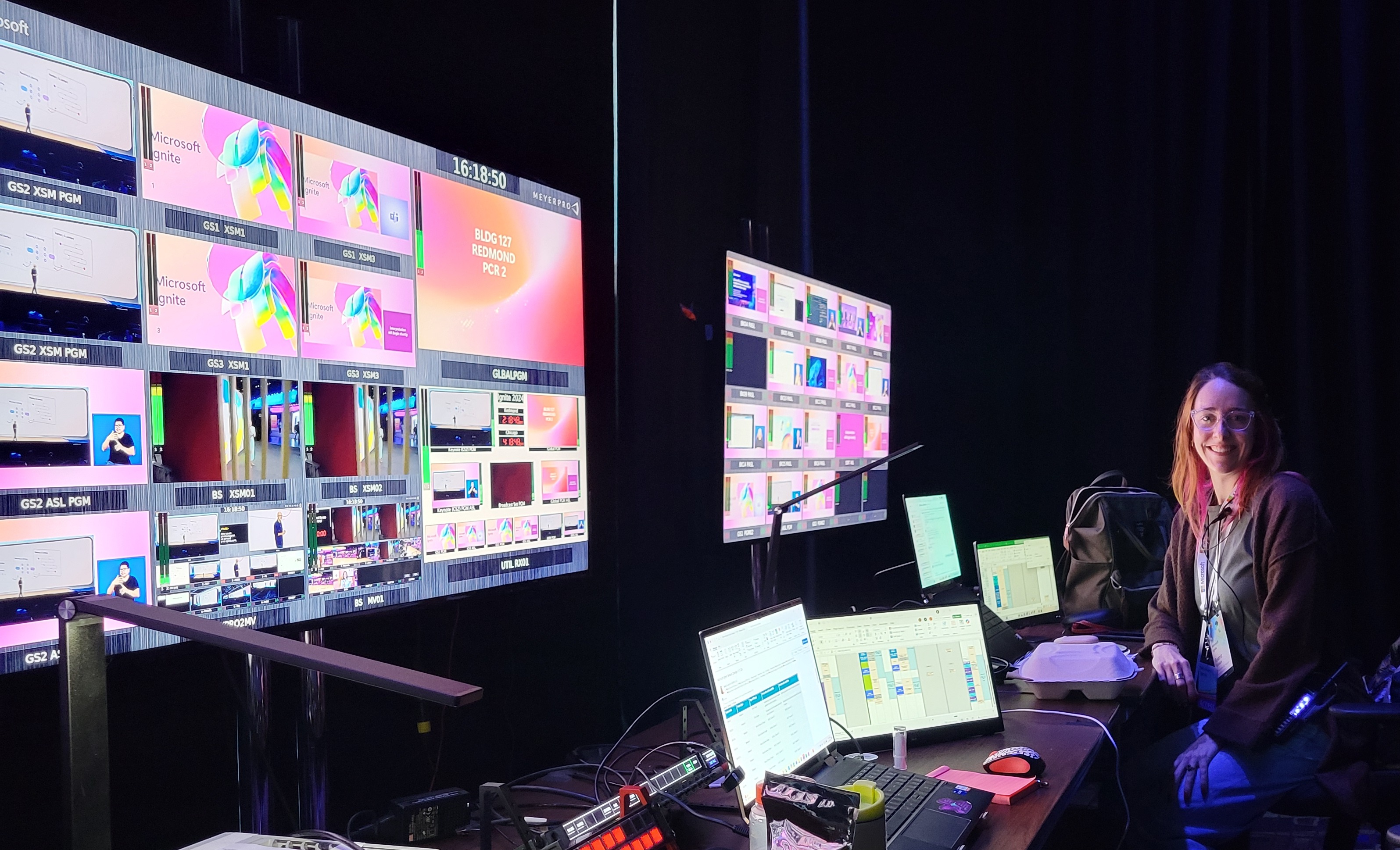 Smiling person with glasses at desk with laptops and multiple large screens showing Microsoft Ignite content.