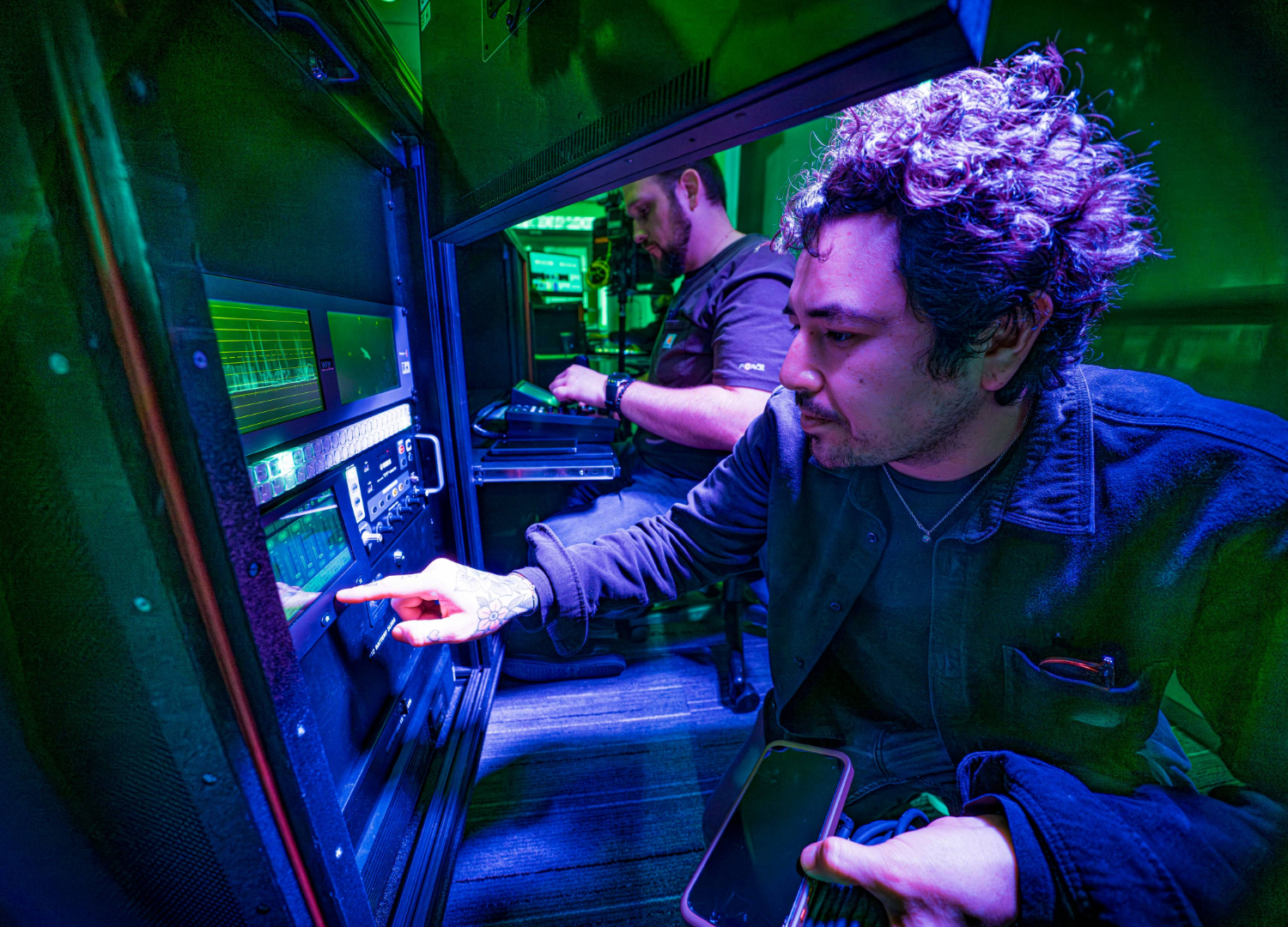 Two men in a dimly lit tech room working with control panels and electronic equipment.