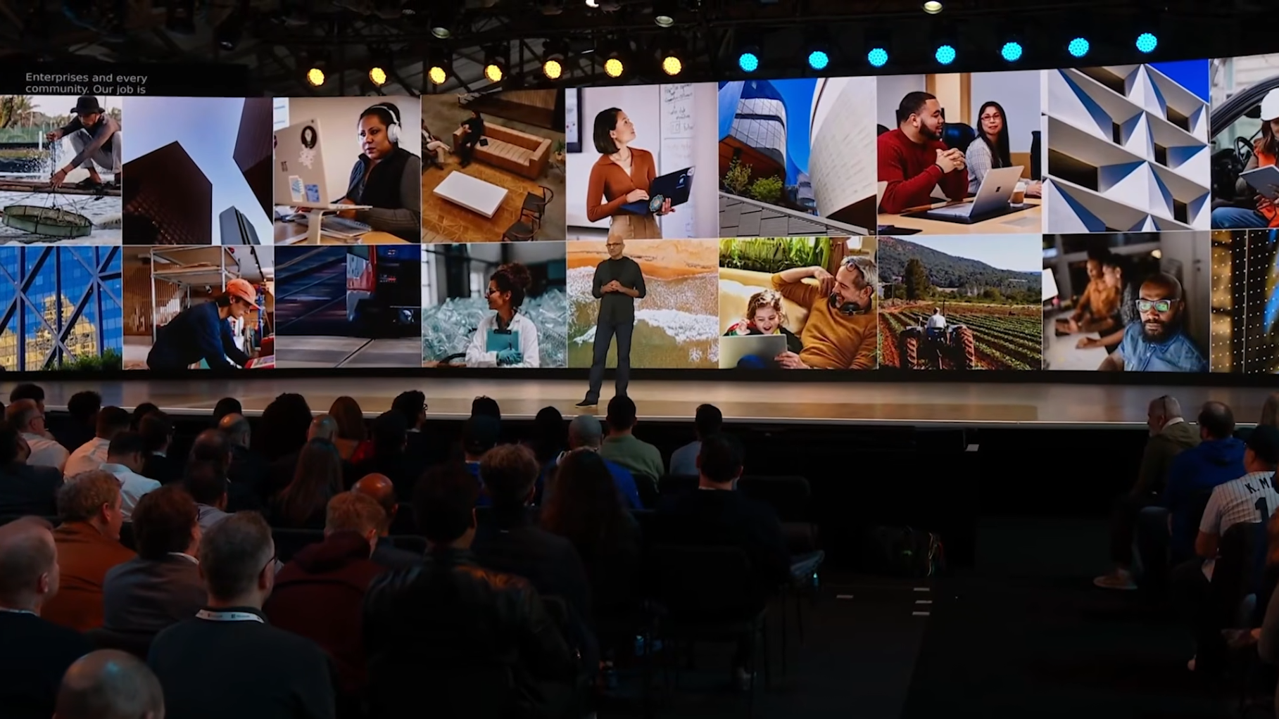 Man presenting on stage to an audience with large screen displaying diverse work and life scenes behind him.