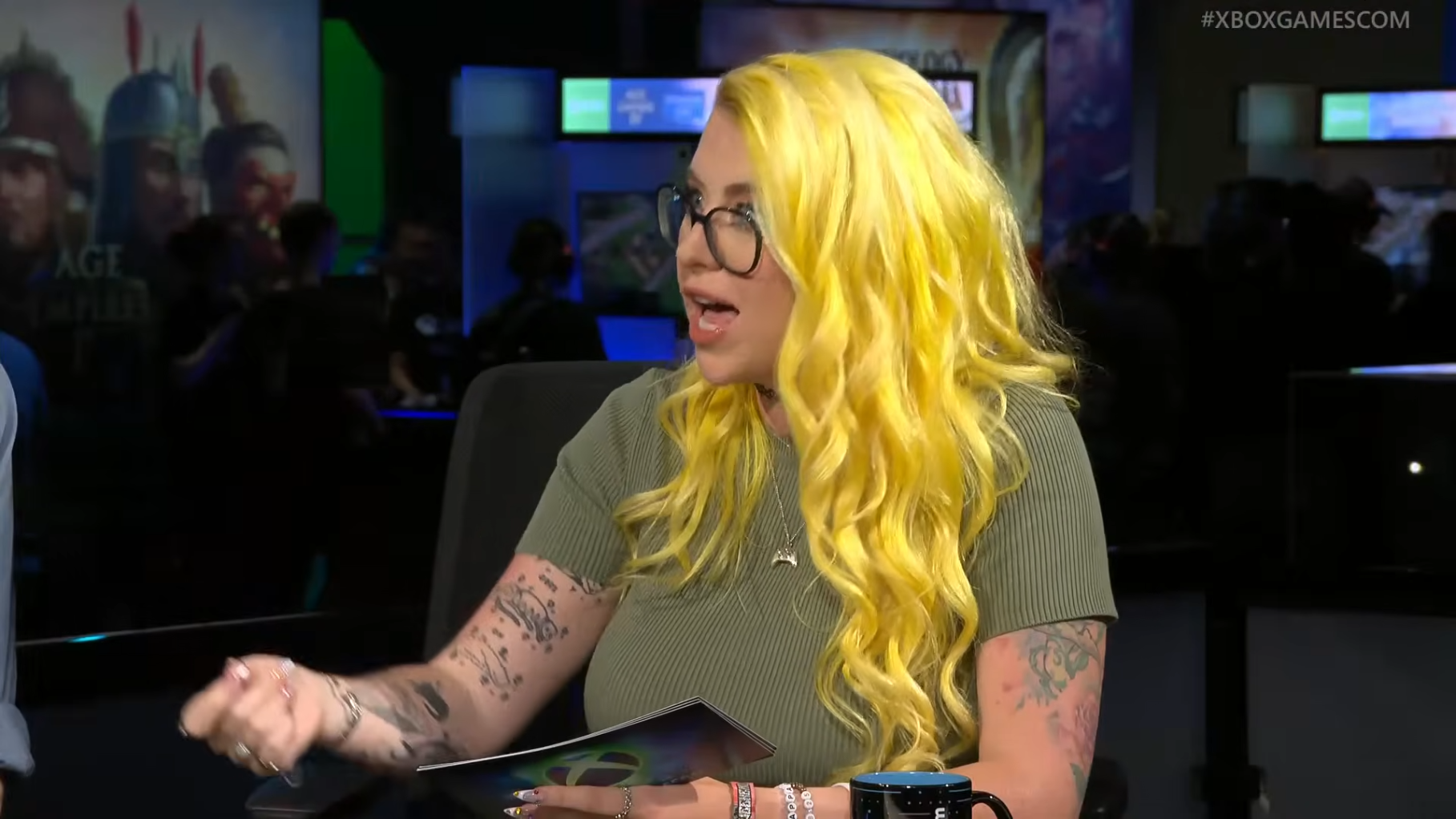 Woman with long yellow hair and glasses speaking while holding papers at a gaming event.