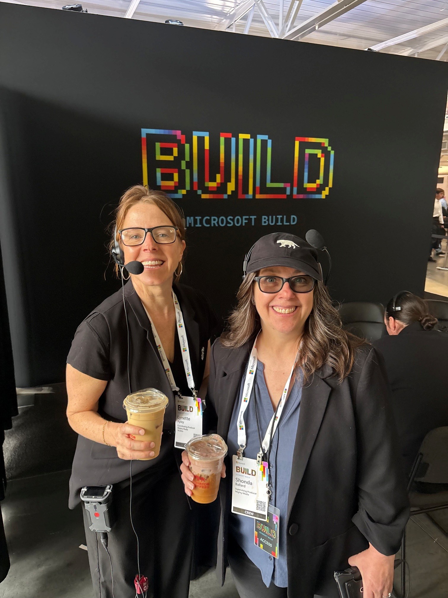 Two women with headsets holding iced drinks in front of a black wall with colorful BUILD Microsoft text.