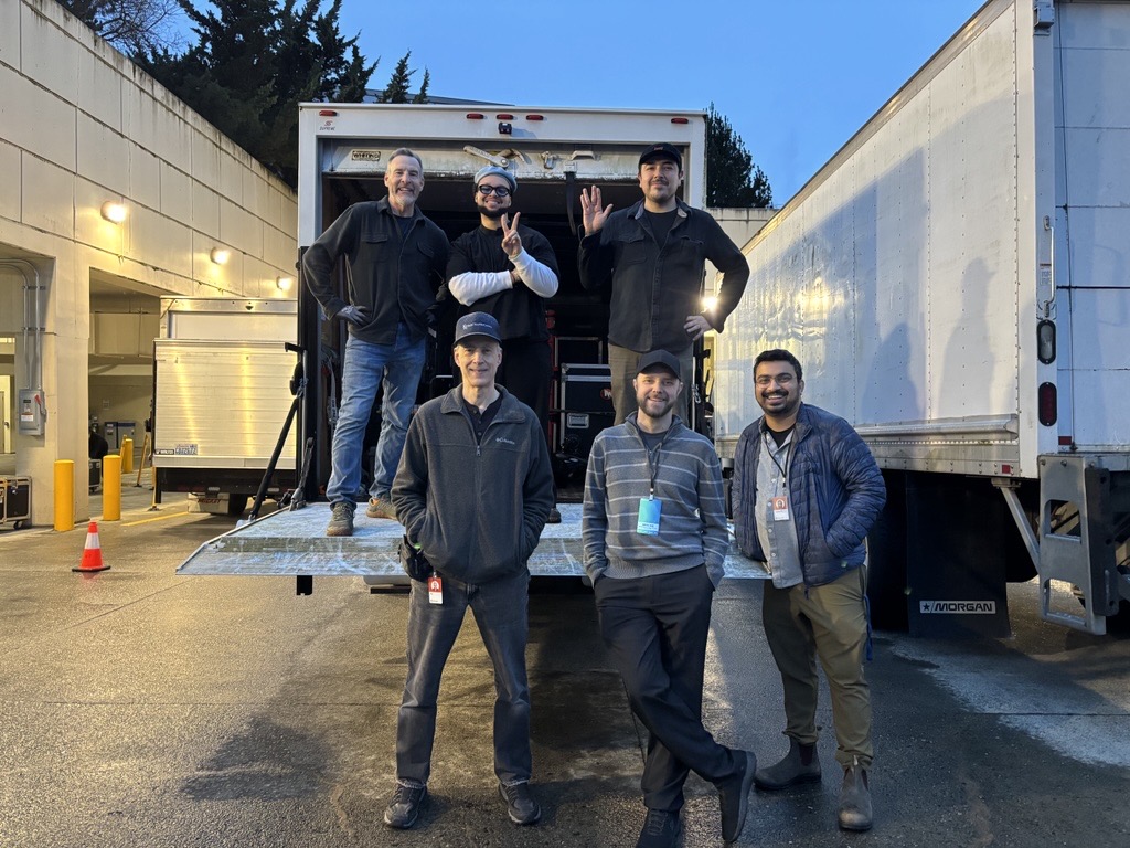 Six men smiling and posing on and in back of an open delivery truck at dusk.