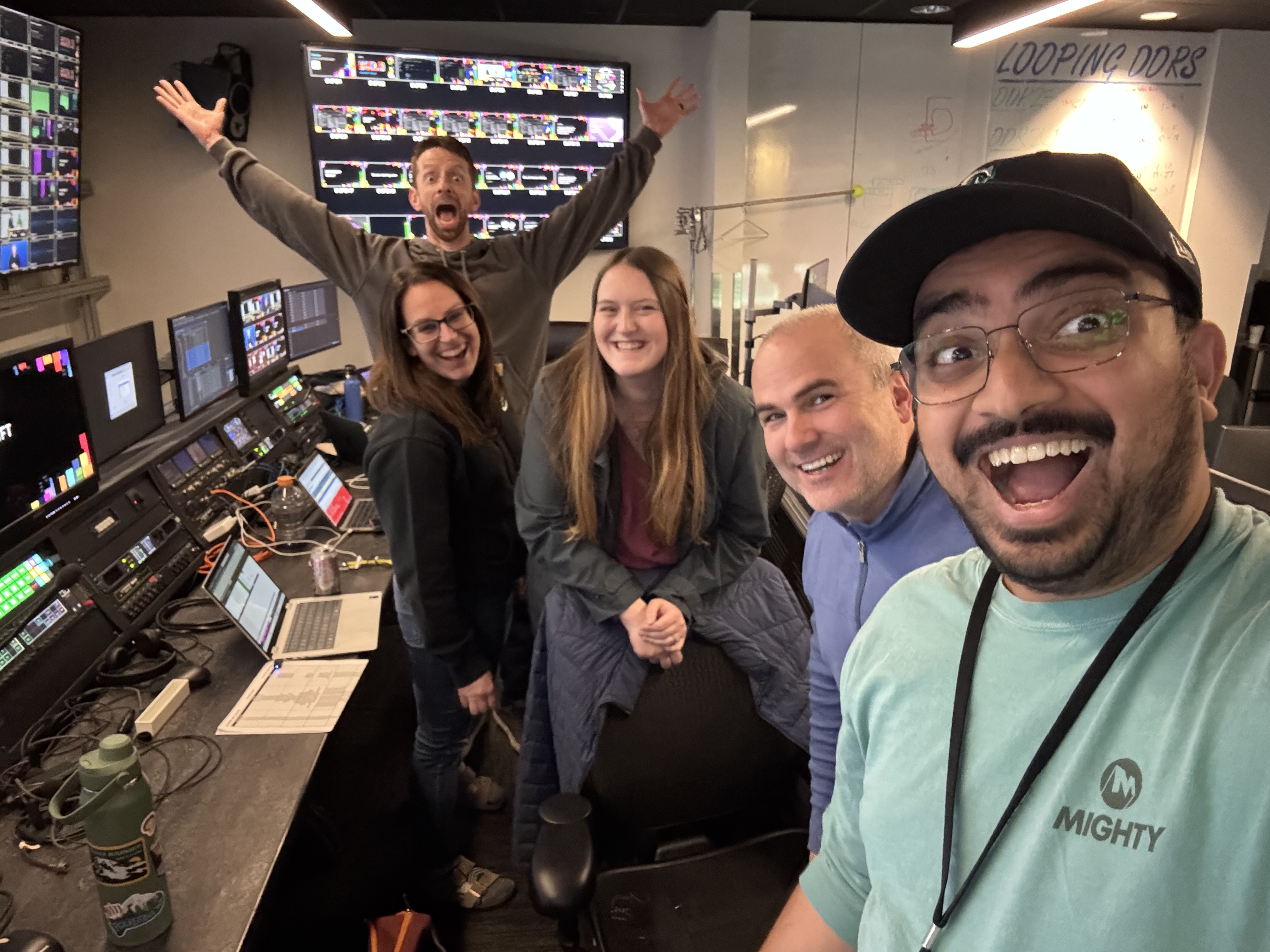Five smiling people in a control room with multiple monitors and equipment, one raising arms excitedly.