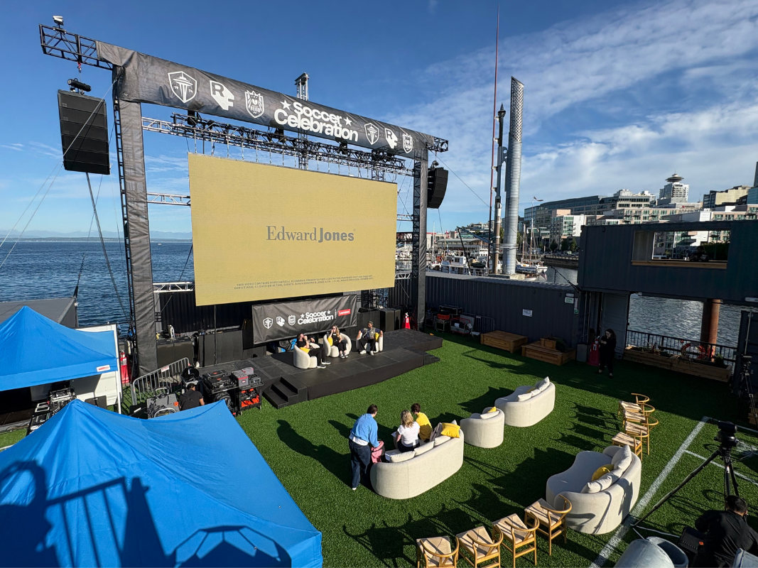 Outdoor stage with large screen and people sitting on cream sofas on artificial grass by the waterfront.