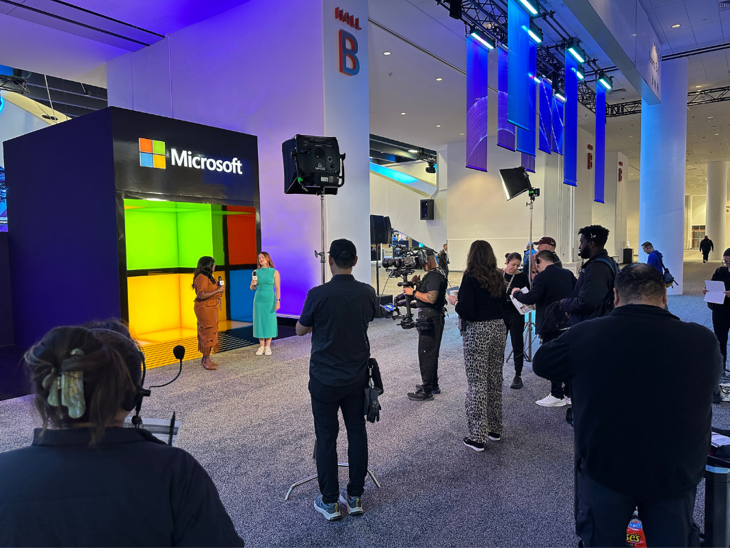 Two women speaking with microphones beside an illuminated Microsoft display at an indoor event with a camera crew.