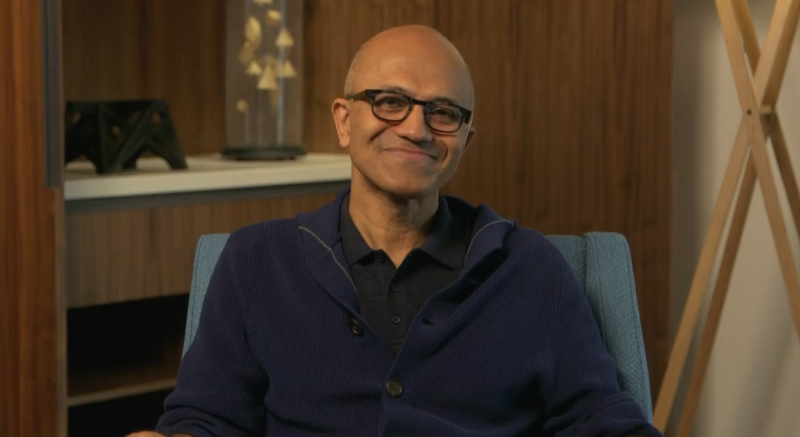 Smiling man with glasses wearing a navy sweater seated in a modern room with wooden decor.