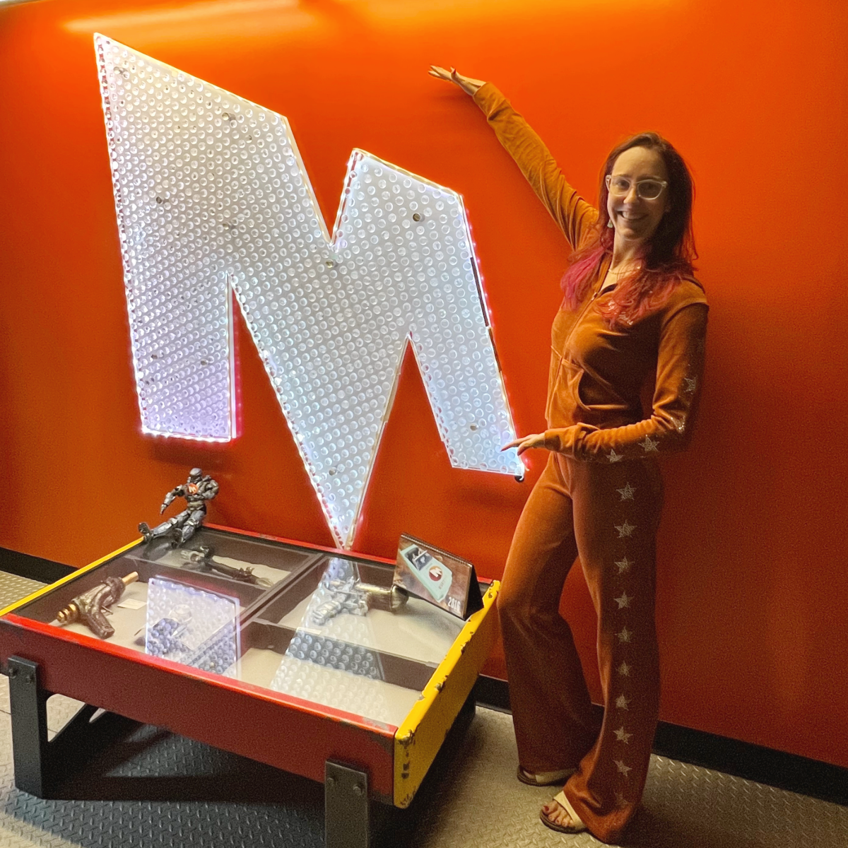 Smiling woman in orange outfit gestures at large lit letter M on orange wall above display case.