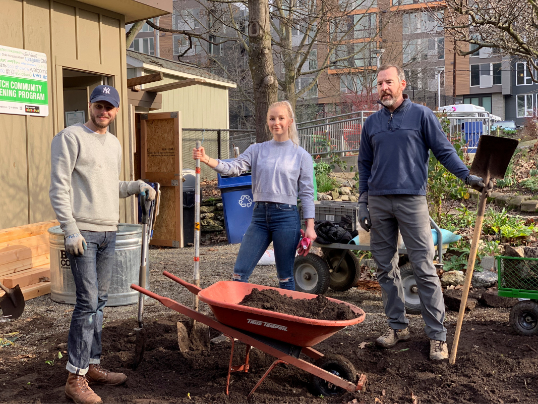 Three people with shovels and a wheelbarrow filled with soil in a garden near a community building.