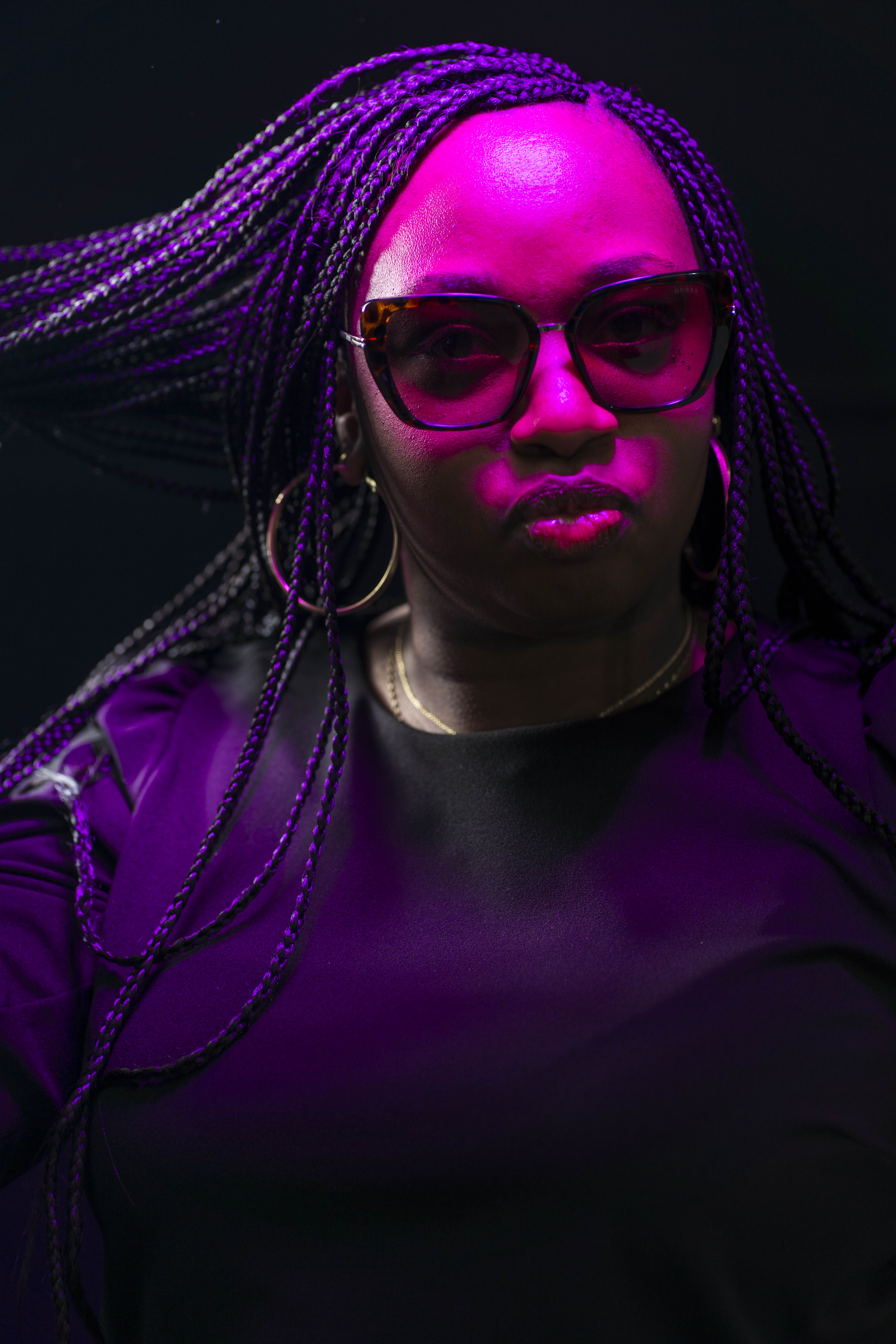 Woman with long braided hair wearing large sunglasses and hoop earrings under purple lighting.