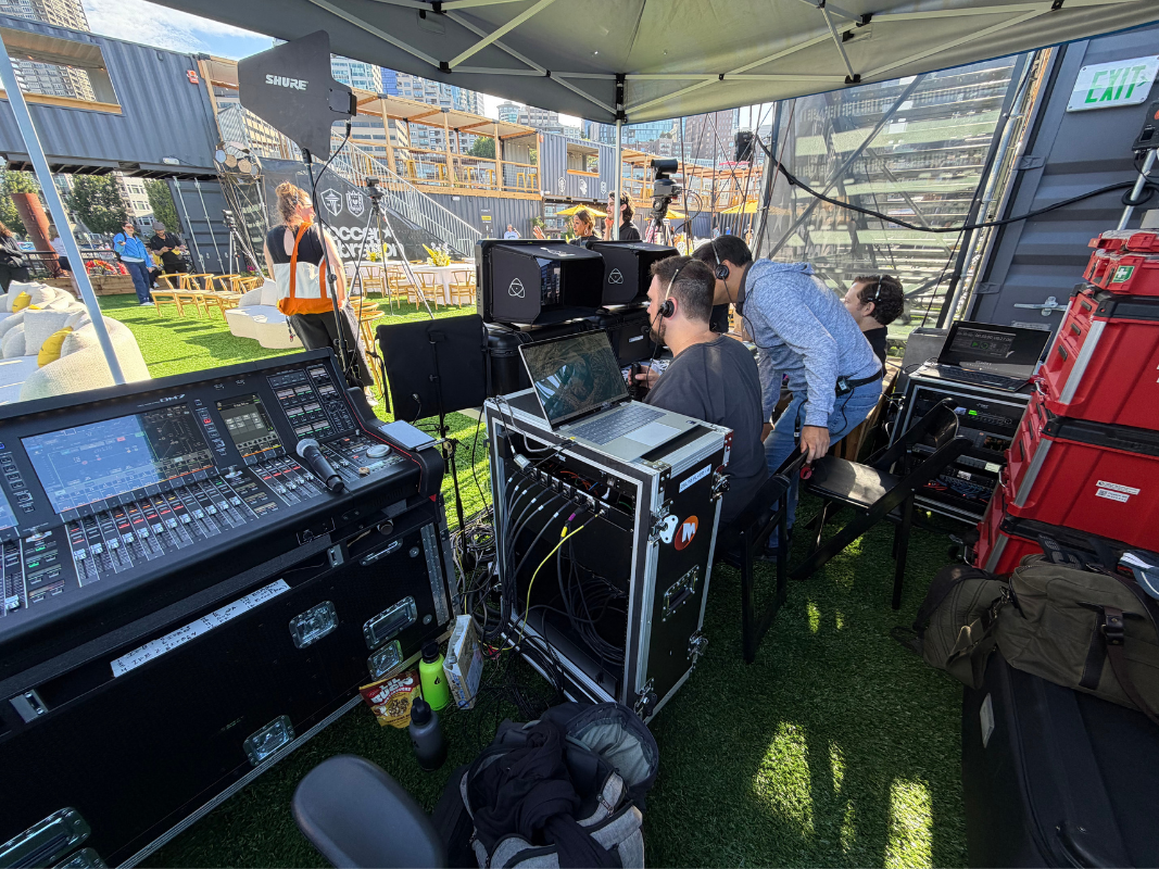 Outdoor video production crew operating equipment and computers under a canopy on artificial grass.
