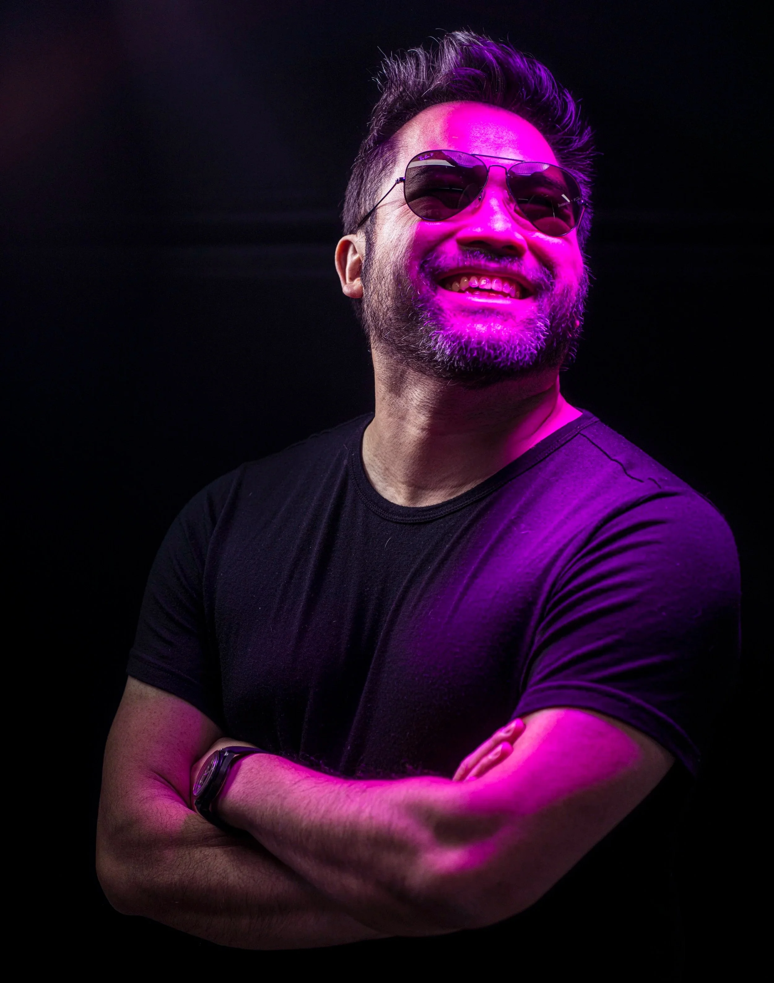 Man with beard and sunglasses smiling, arms crossed, lit with purple light on black background.