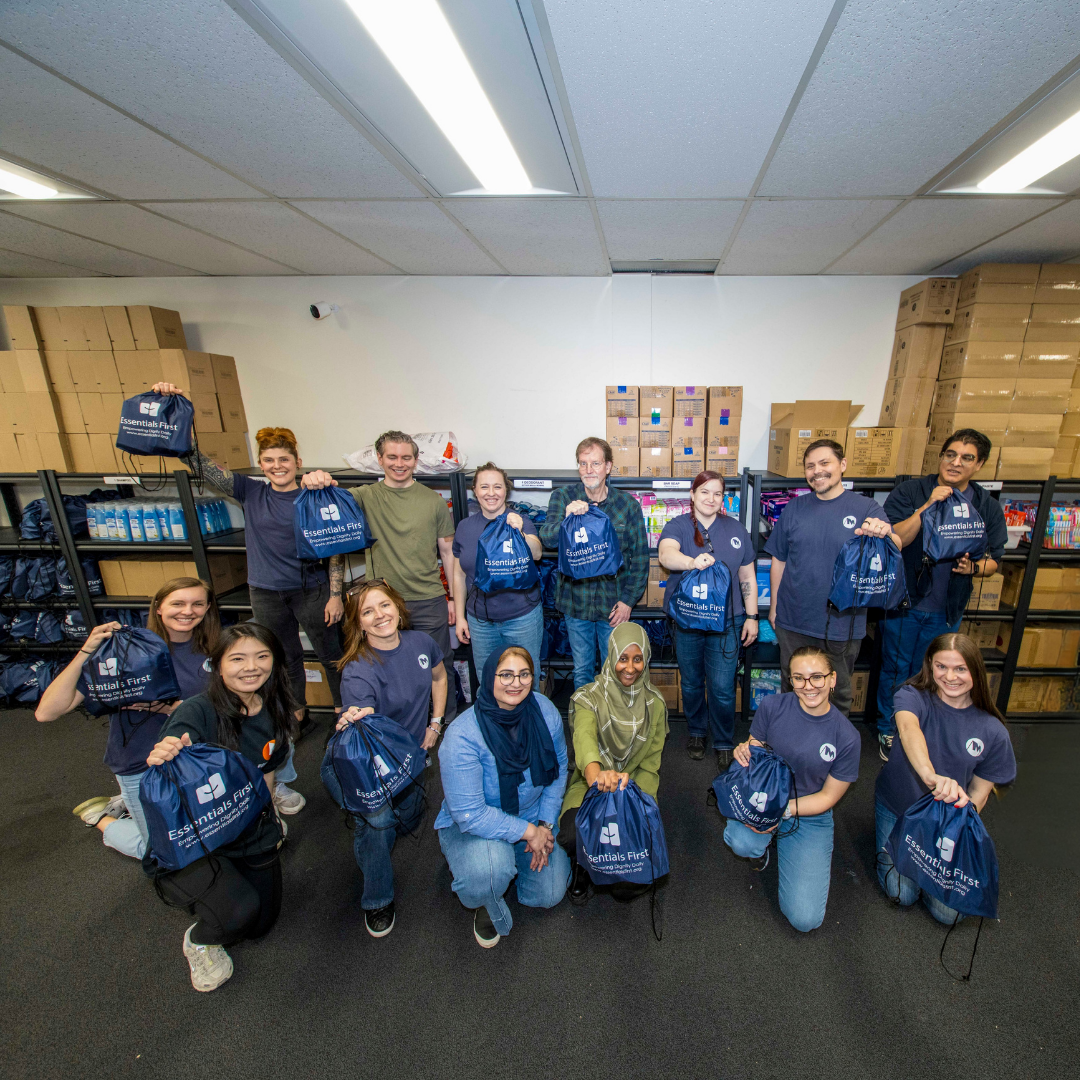 Group of volunteers holding blue bags labeled Essentials First in a room with shelves and boxes.