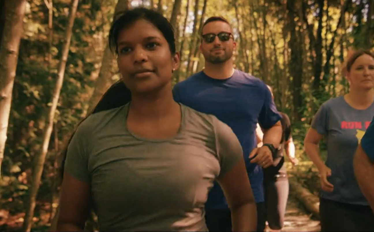 People jogging on a forest trail with sunlight filtering through the trees.