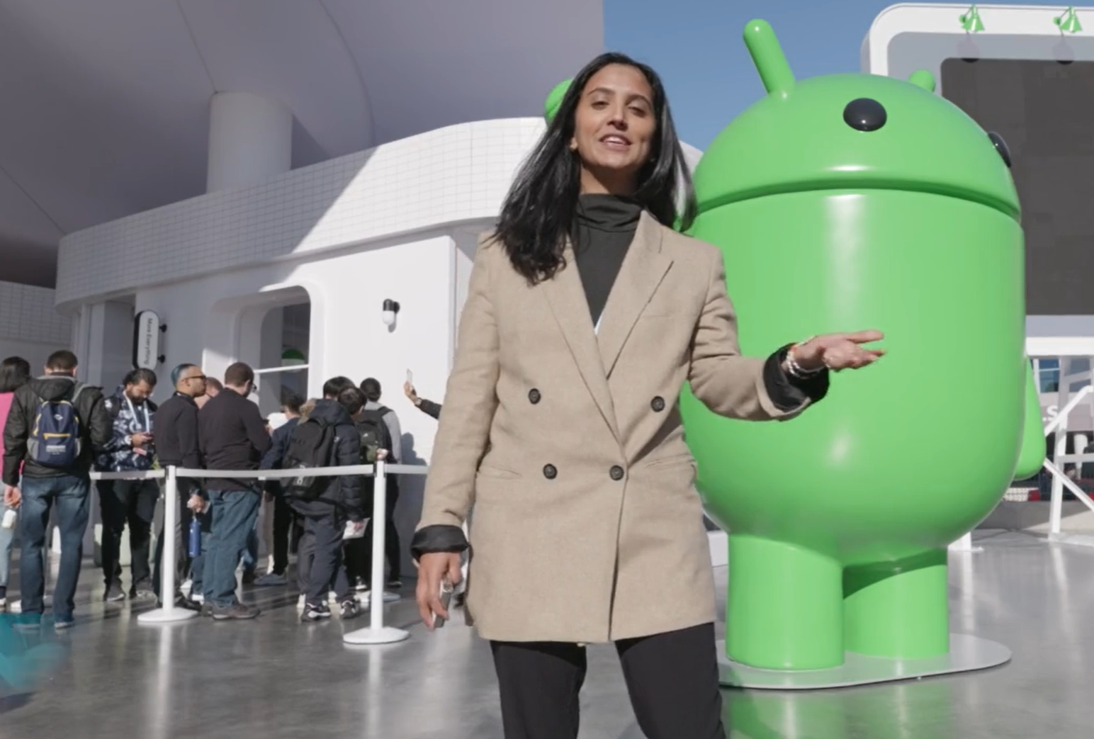Woman in beige coat speaking with large green Android statue and people queuing in background.