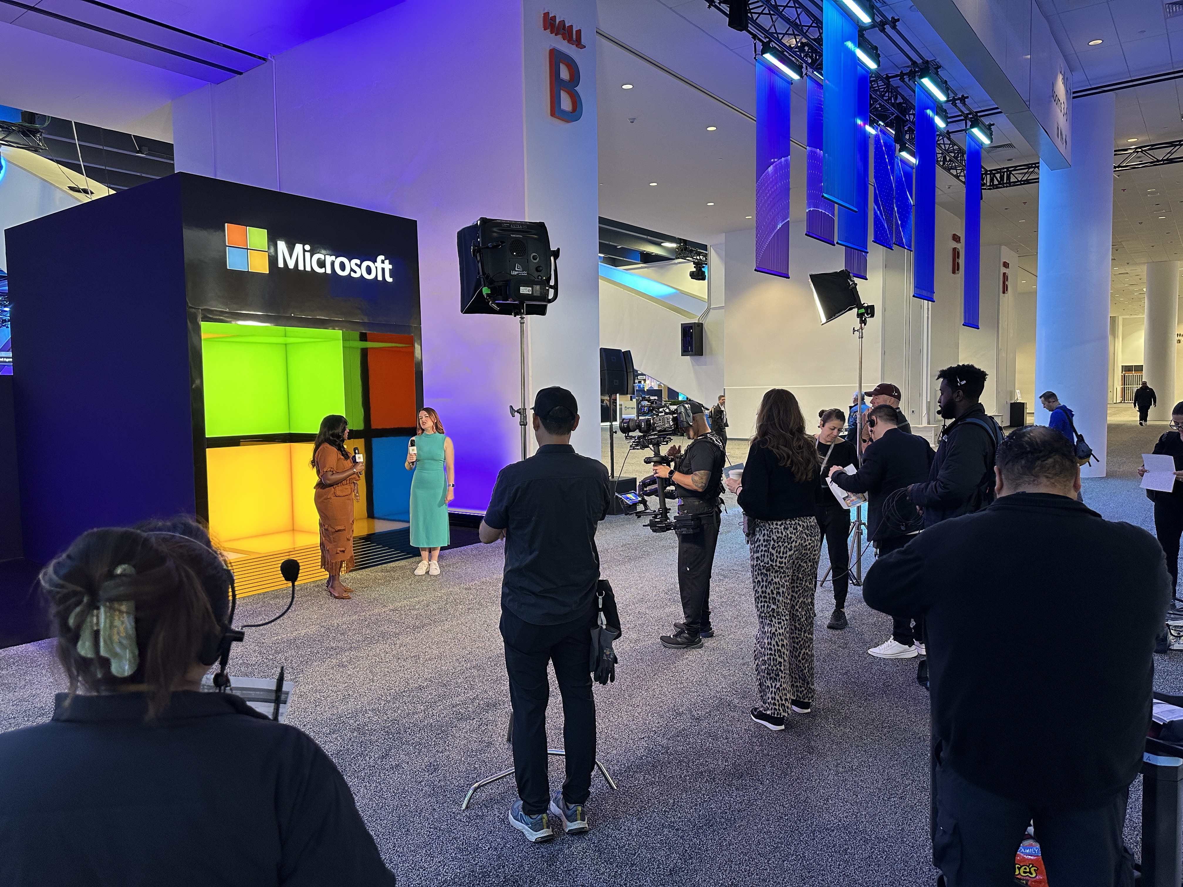Video crew records two women speaking in front of a large illuminated Microsoft logo display.