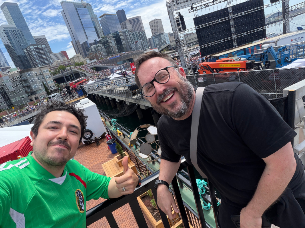 Two men smiling at the camera on a balcony with a city skyline and a crowd behind them.