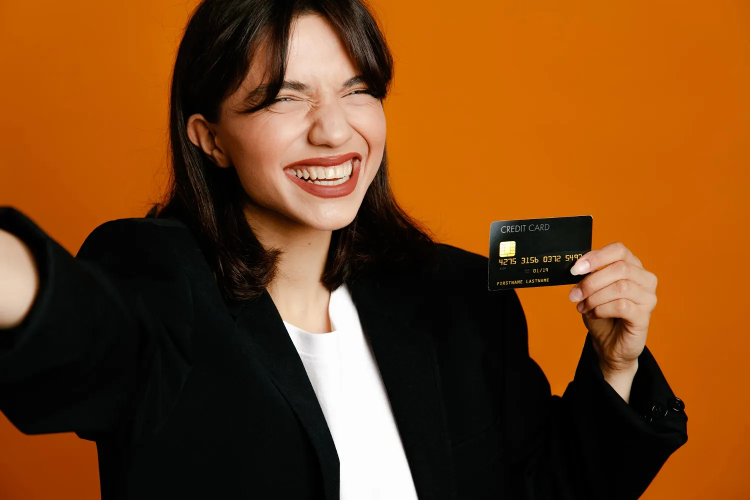 Smiling woman in a blazer holding up a black credit card against an orange background