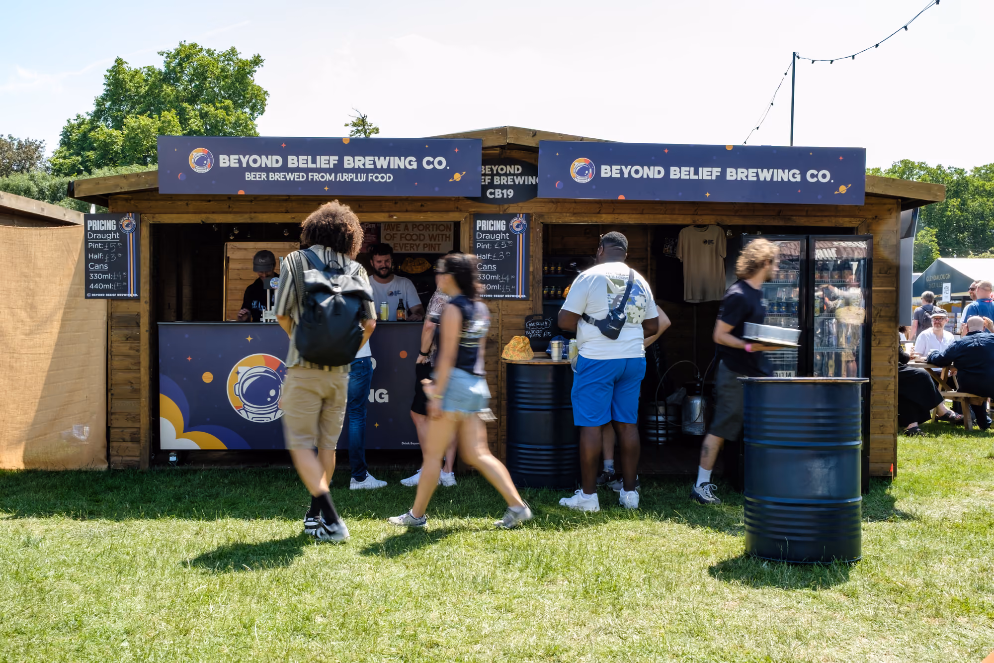 People standing and walking near a wooden Beyond Belief Brewing Co. beer stand at an outdoor event.
