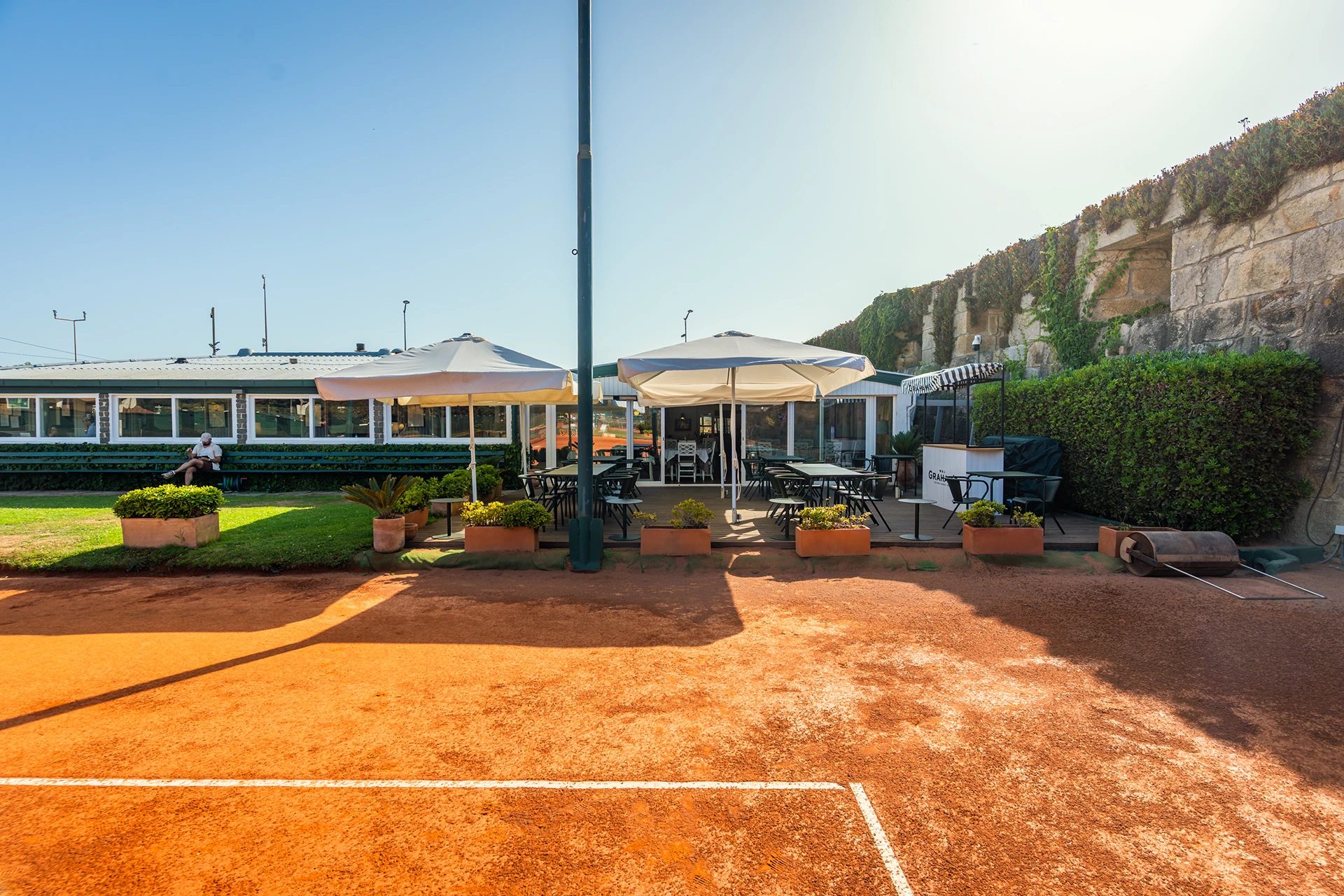 Outdoor seating area with tables, umbrellas, and plants beside a clay tennis court and building.