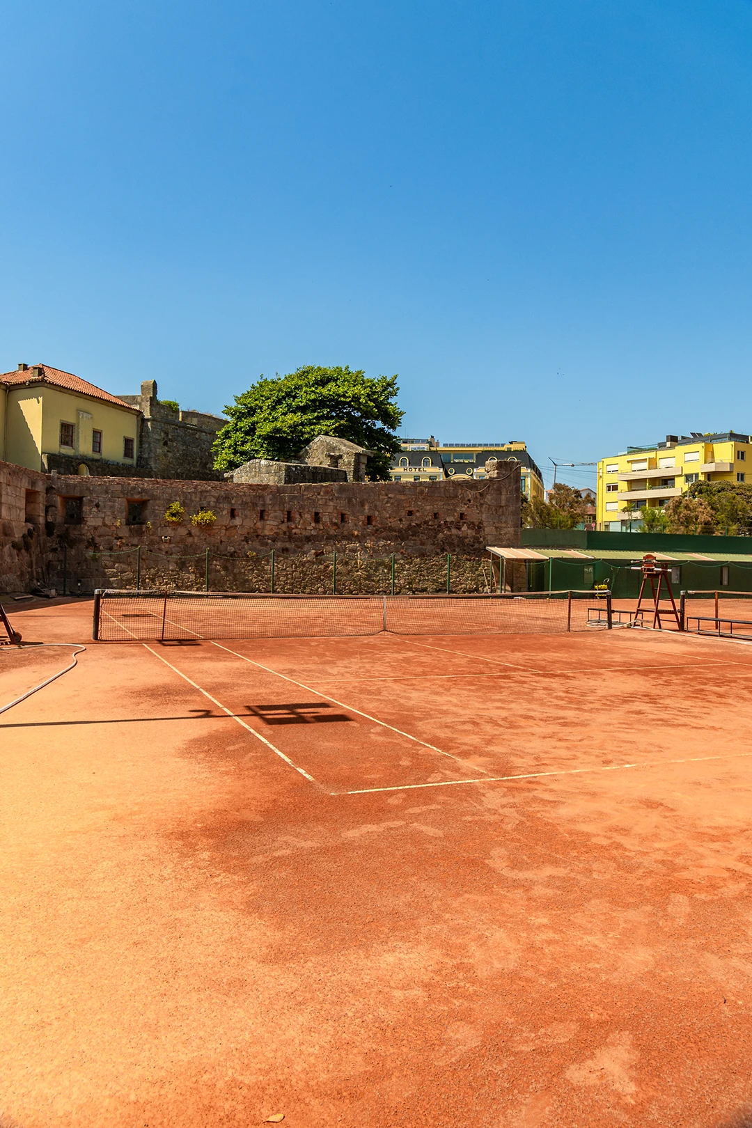 Empty clay tennis court with net, surrounded by stone walls and buildings under clear sky.