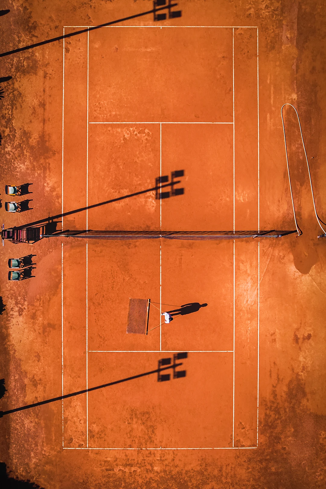 Aerial view of a clay tennis court with one person and shadows of floodlights and net posts.