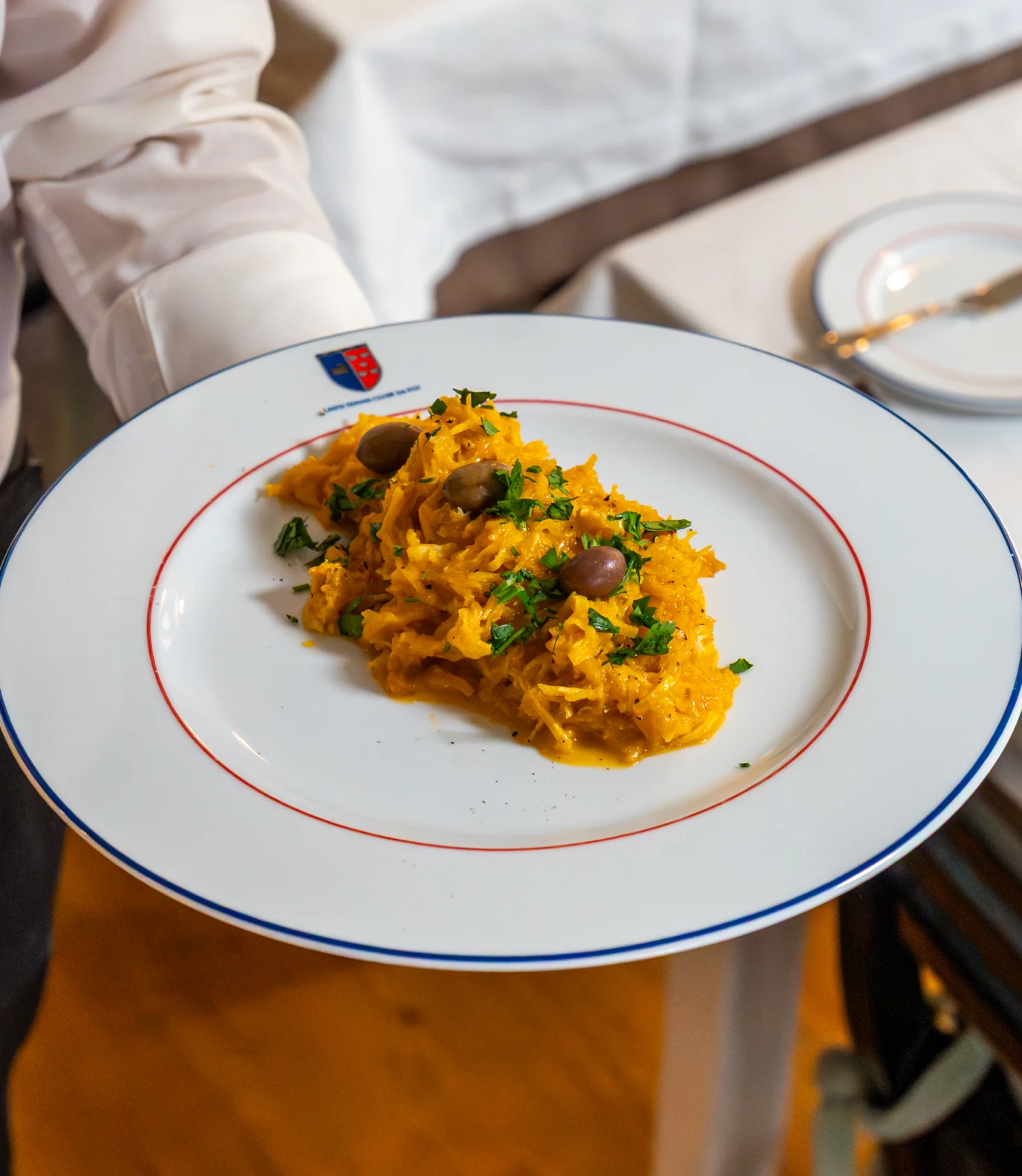 White plate of creamy orange shredded dish garnished with olives and chopped herbs held by a person.