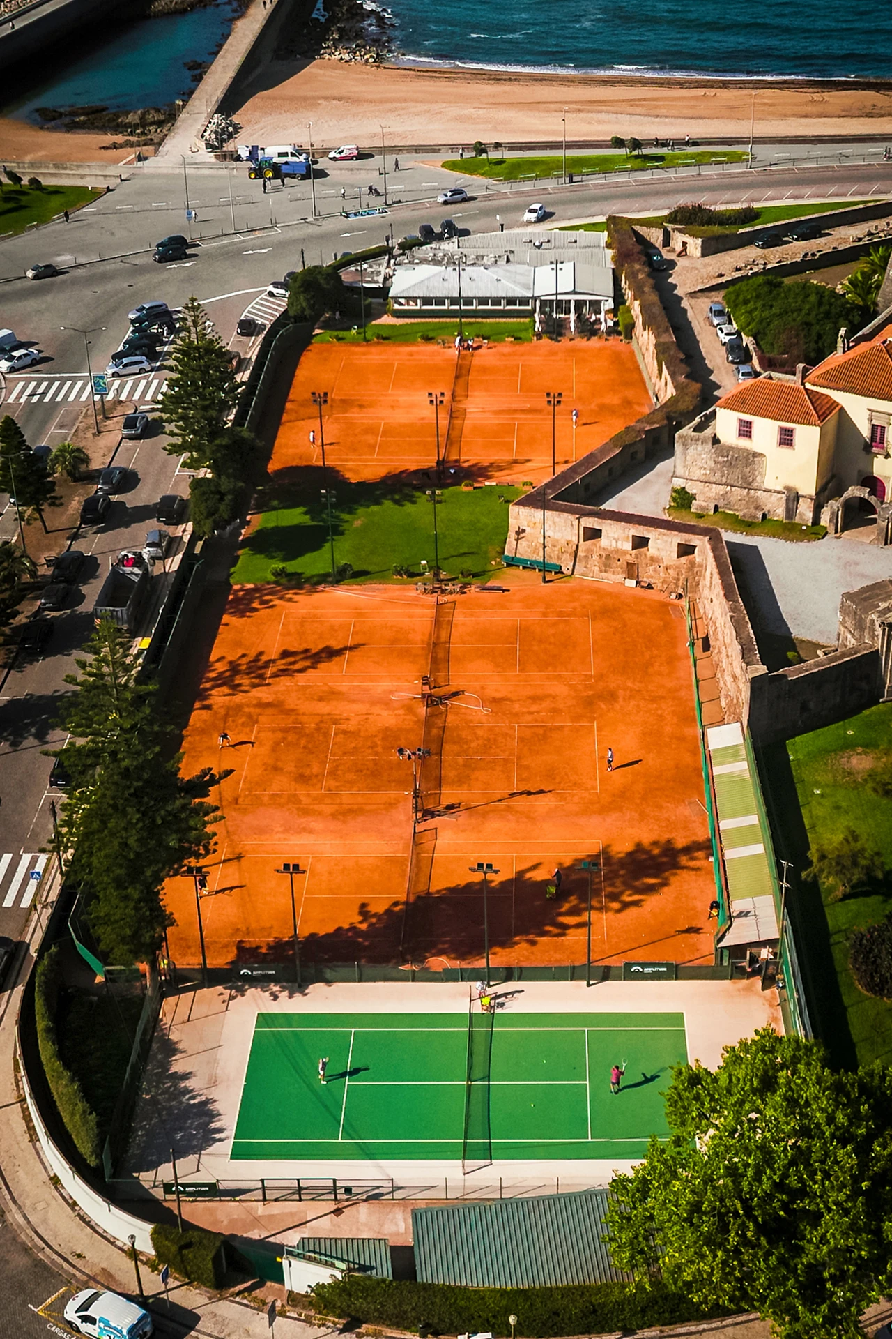 Aerial view of clay and hard tennis courts near a beach with players and surrounding buildings.