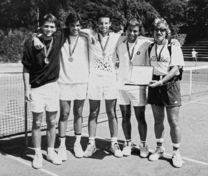 Five men standing on tennis court wearing medals, one holding a plaque, posing happily for photo.