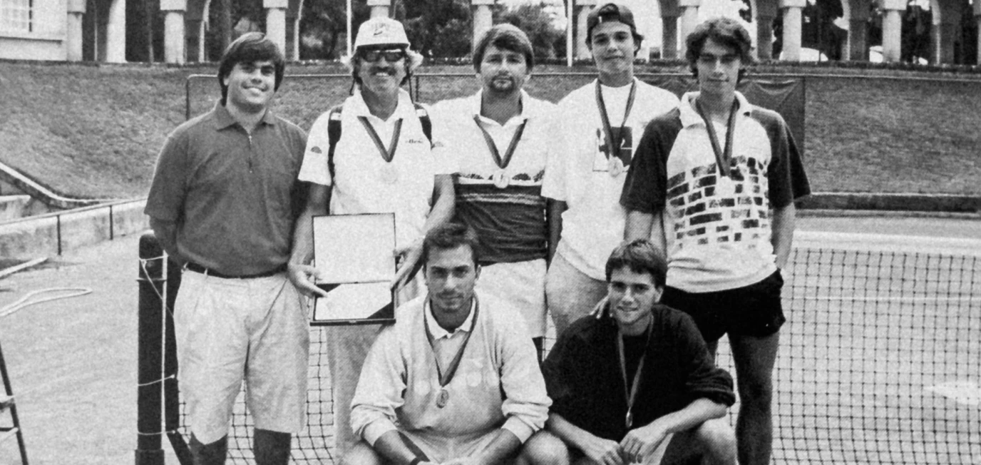 Seven men posing on a tennis court, most wearing medals, one holding a plaque.