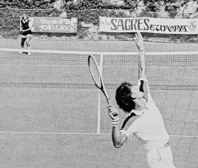 Black and white photo of a male tennis player serving on an outdoor court with banners in the background.