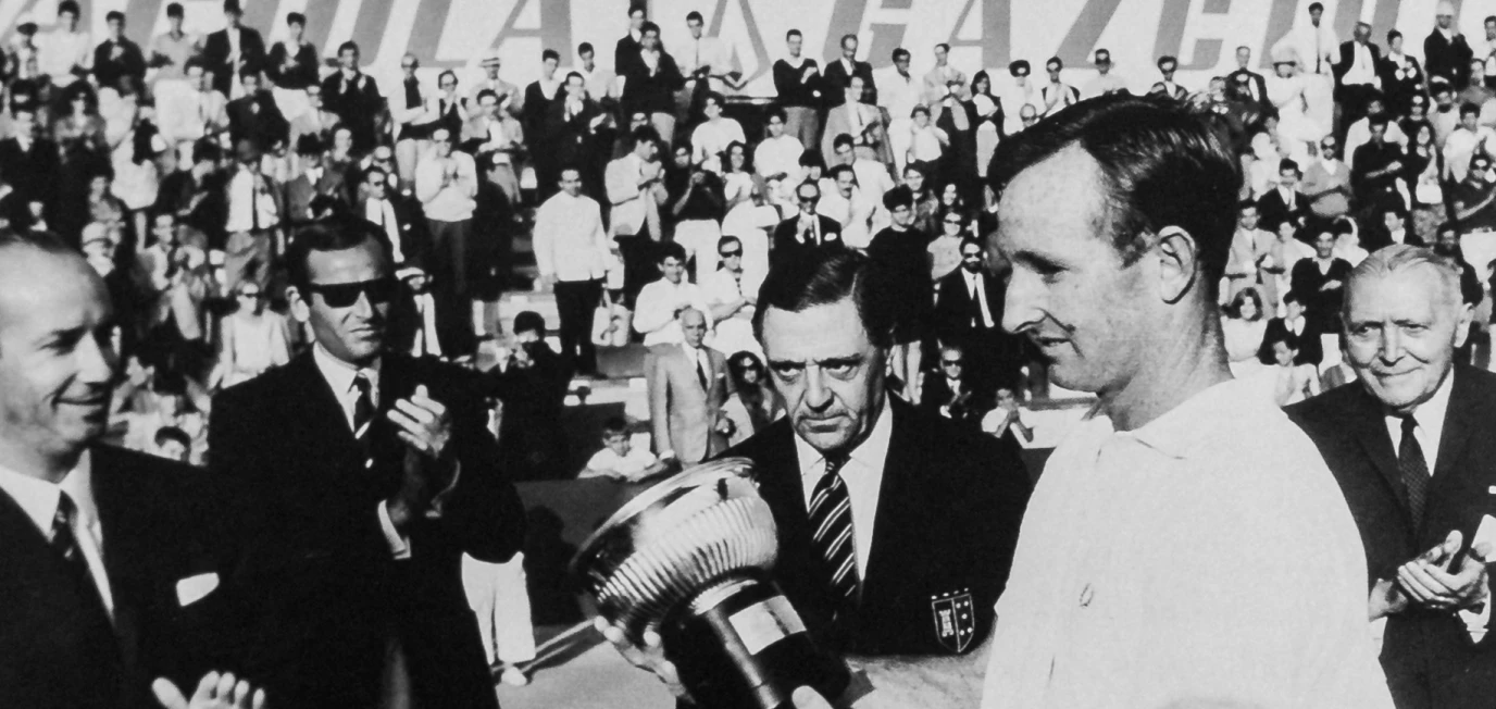 Black and white photo of a man in white receiving a trophy from officials with people clapping in the background.