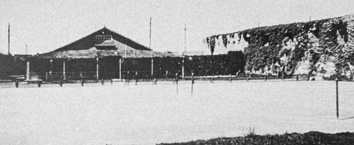 Black and white photo of a large building with a triangular roof behind a fenced area and ivy-covered walls.
