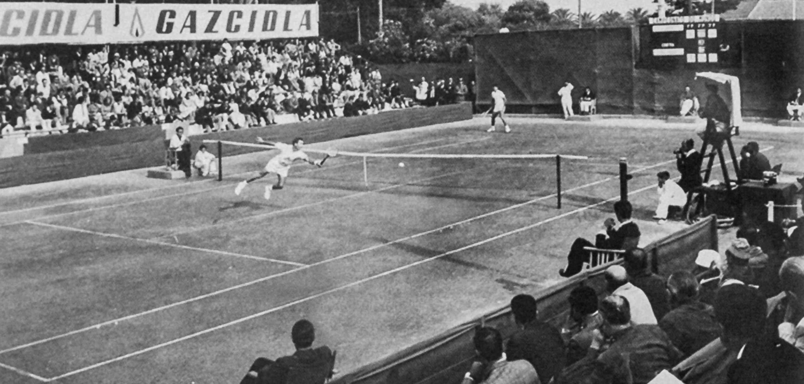 Black and white photo of a tennis match with a player diving for the ball and a crowd watching.