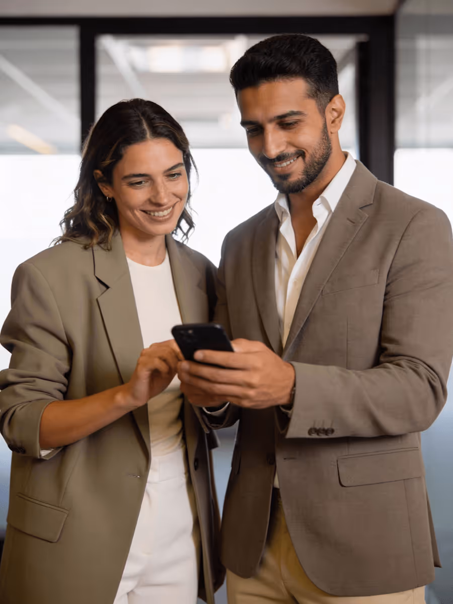 Two professionals in blazers smiling and looking at a smartphone together in an office setting.