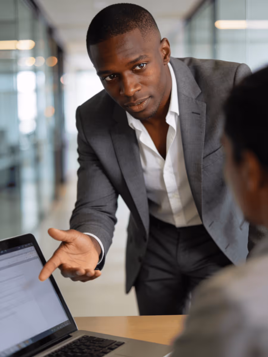 Man in gray suit leaning over desk explaining something to a colleague near a laptop.