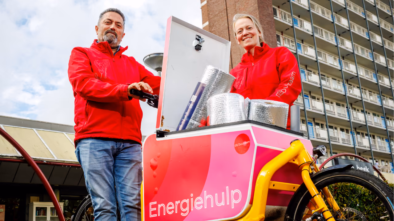 Een mannelijke en vrouwelijke energiehulp poseren voor de camera met hun bakfiets vol met isolatiehulpmiddelen. 