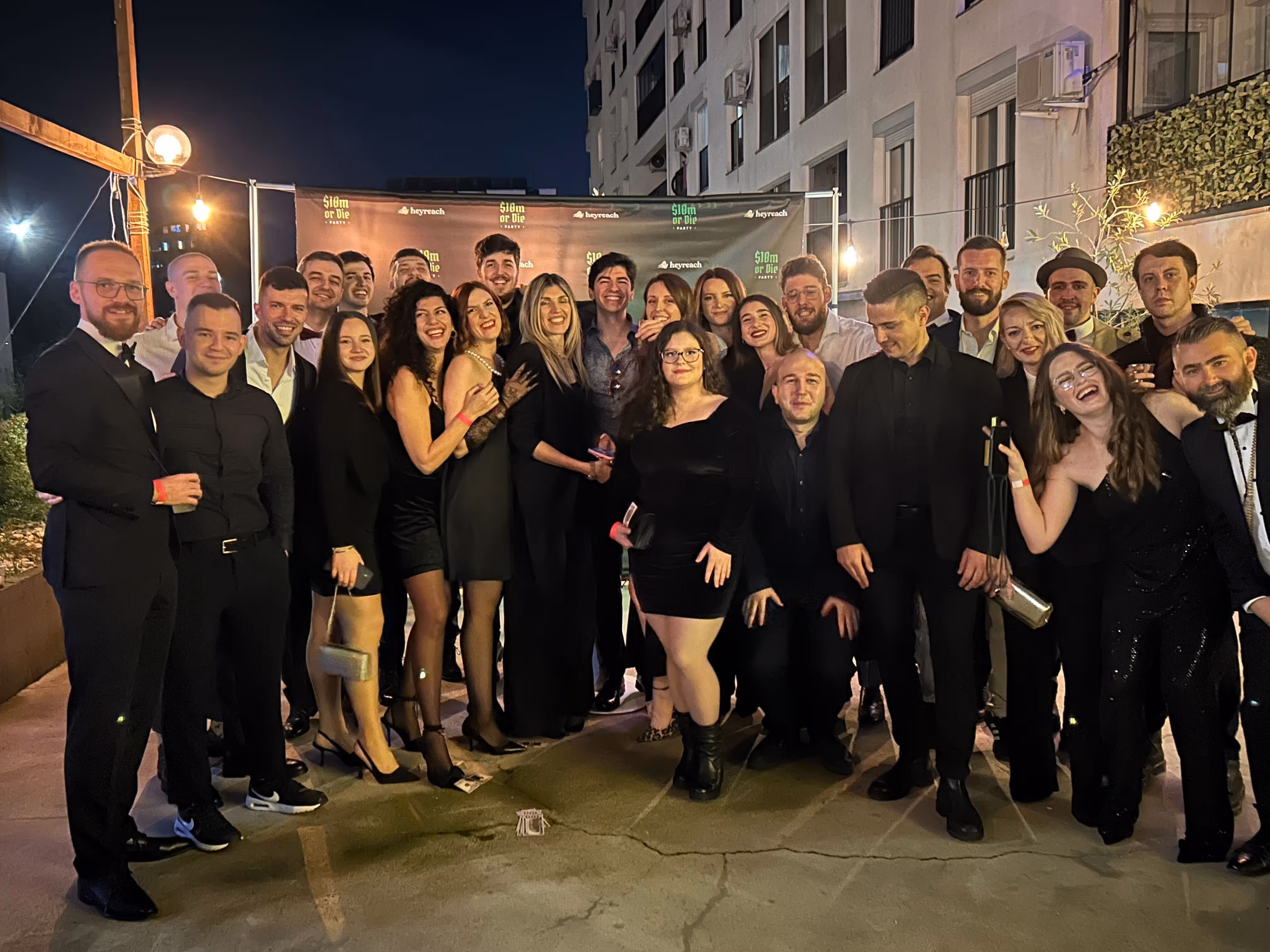 Group of people dressed formally posing and smiling outdoors at night in front of a party banner.