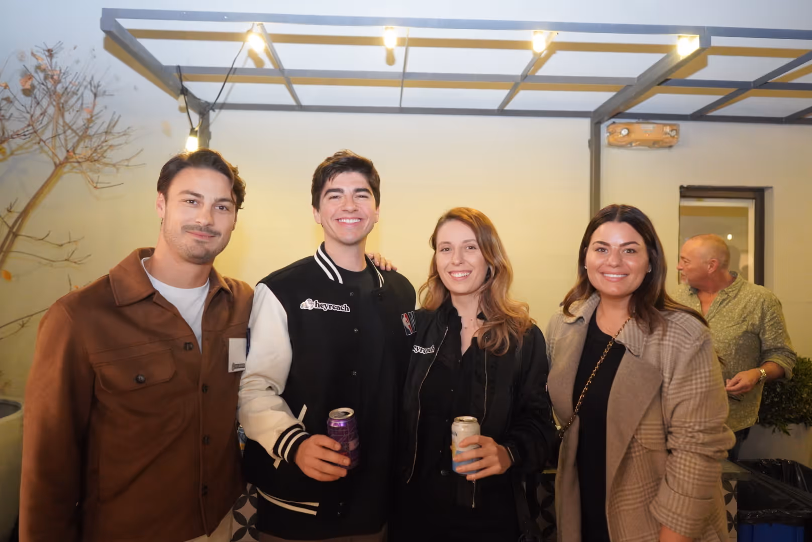 Four people smiling at a social event, with two holding canned drinks under string lights.