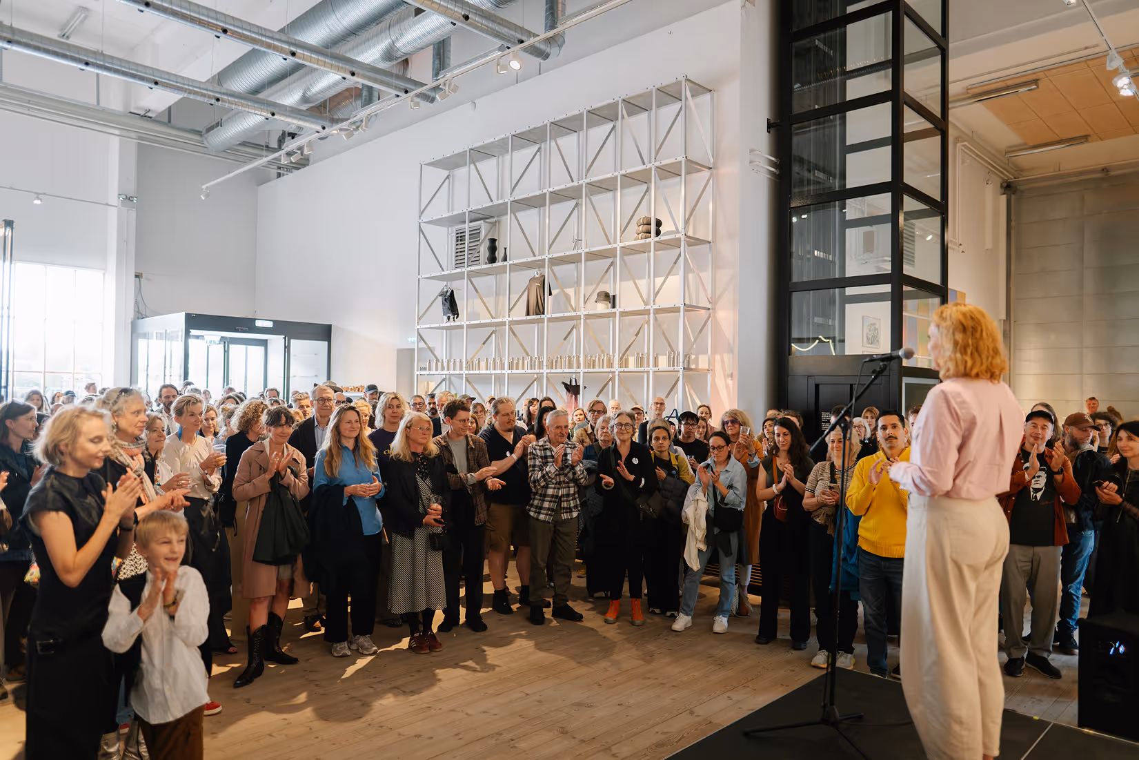 Person speaking on stage to an audience applauding inside a bright modern gallery space.