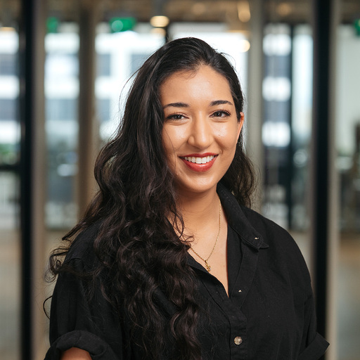 Smiling woman with long curly hair wearing a black shirt in a modern office setting.