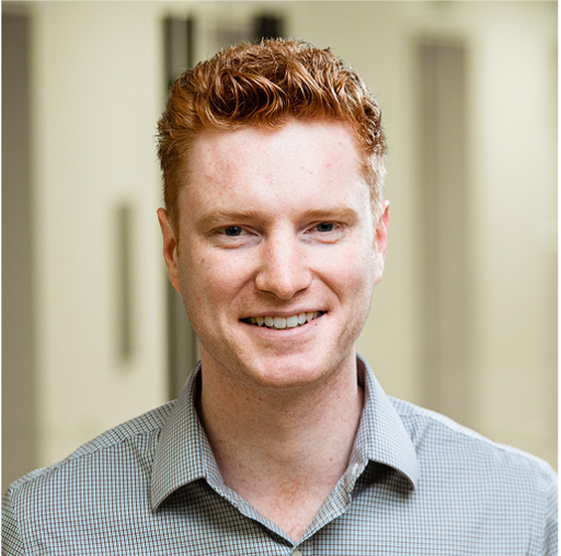 Smiling young man with red hair wearing a checkered shirt against a blurred indoor background.