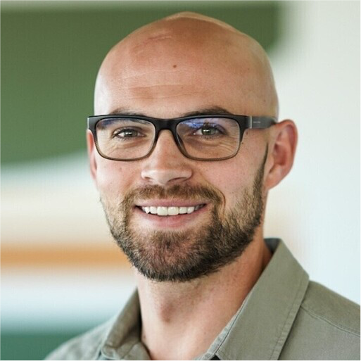 Smiling bald man with glasses and a beard wearing a green shirt, with a blurred background.