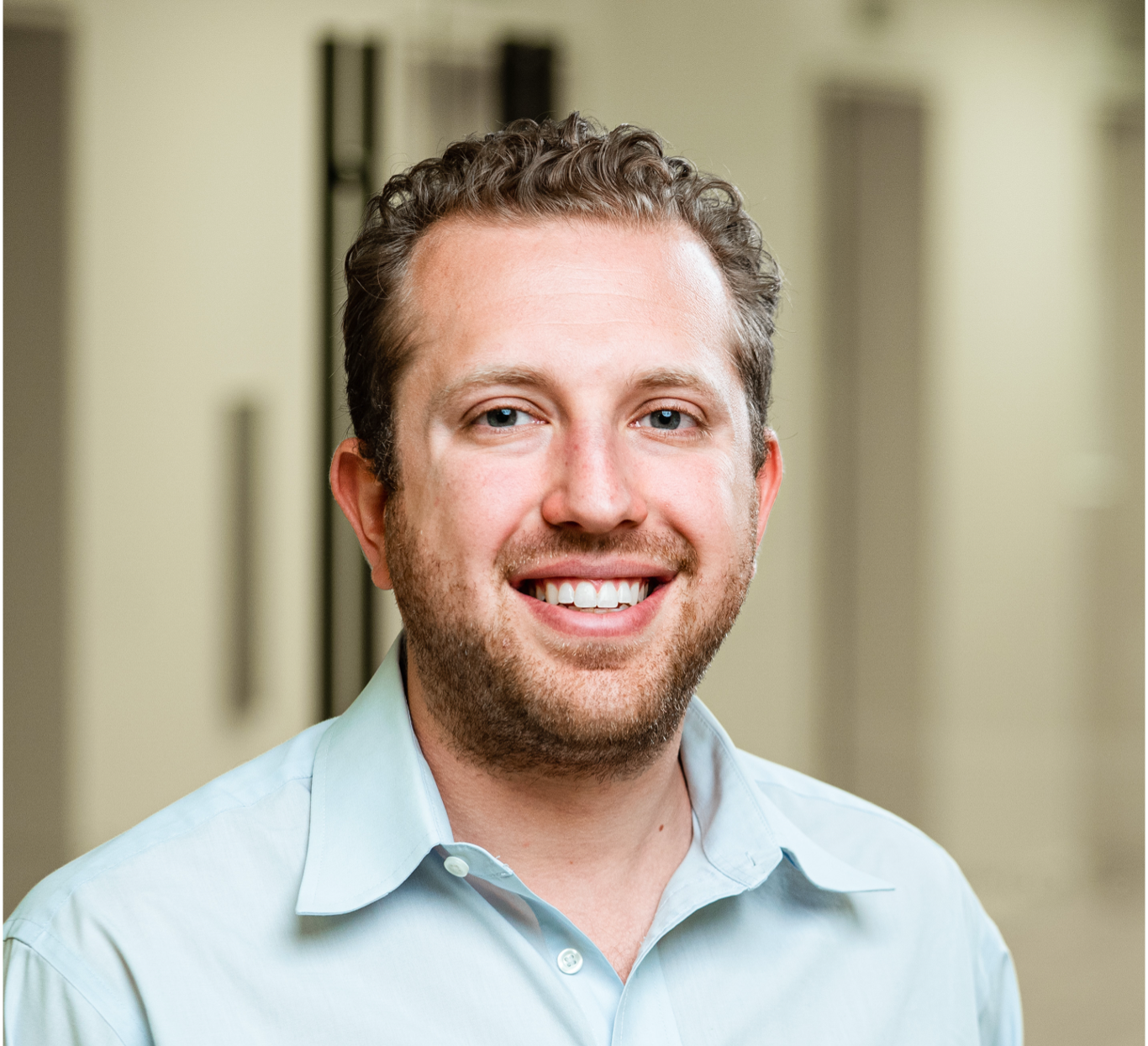 Smiling man with curly hair and beard wearing a light blue shirt in a softly blurred indoor setting.